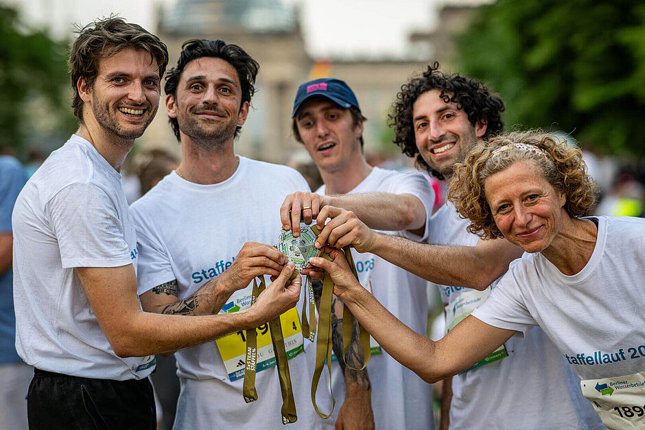 5 glückliche Teilnehmende setzen ihre Medaillen zur runden Teammedaille zusammen und halten sie in die Kamera.